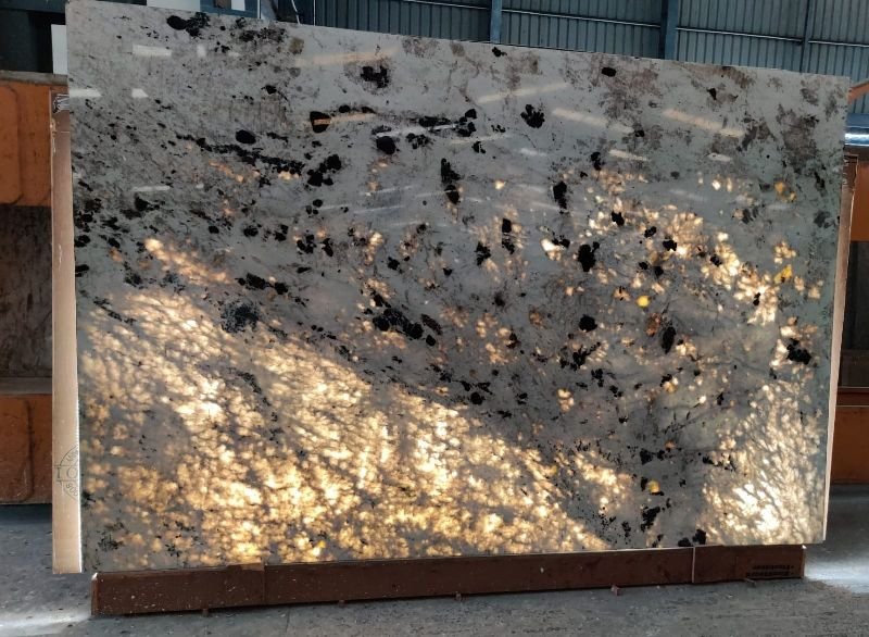 Dalle de granit blanc avec des veines sombres et des amas minéraux noirs, partiellement rétroéclairée pour révéler sa translucidité naturelle